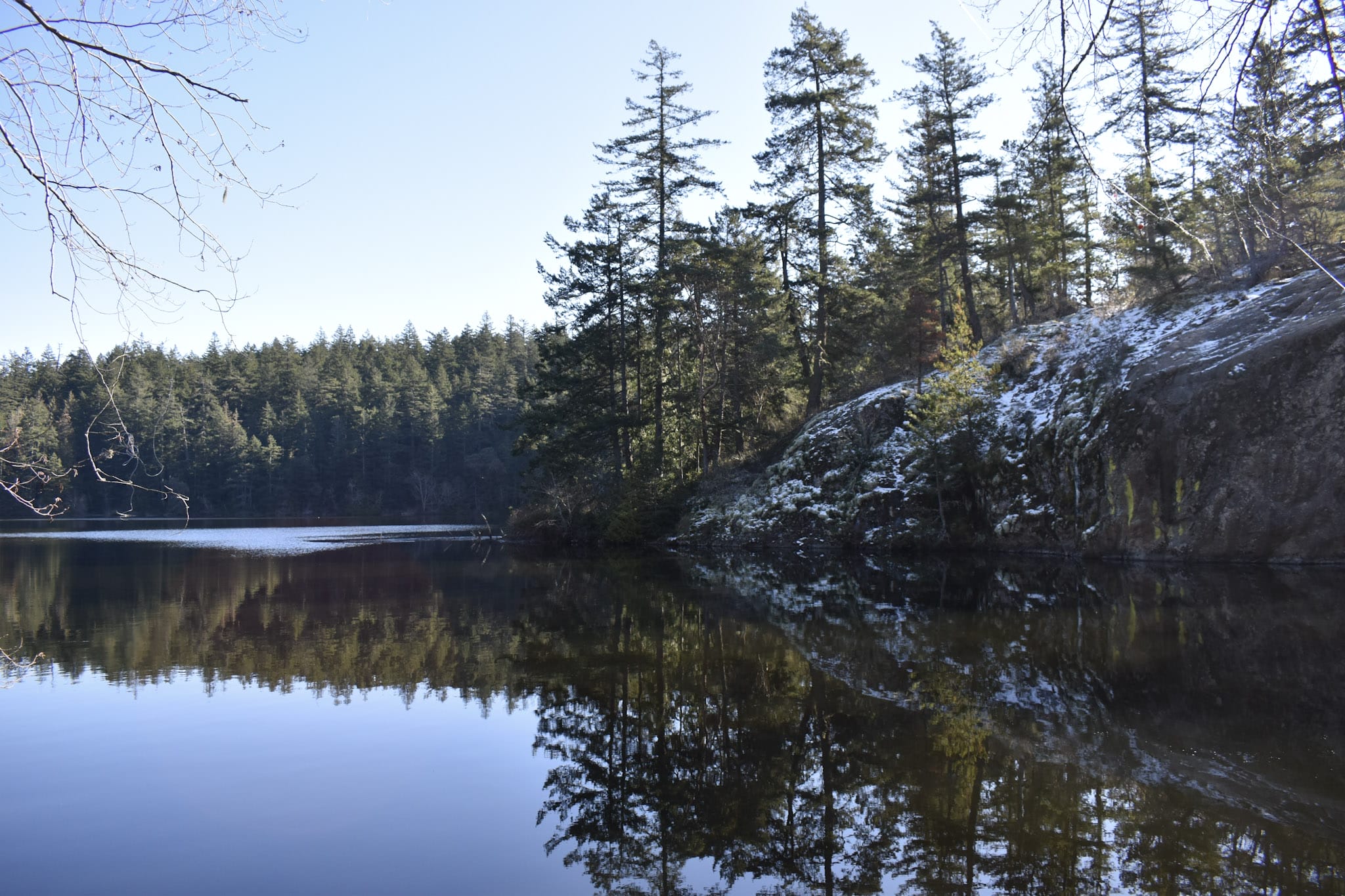 A calm winter lake with evergreen trees and rocks on one side, under a light dusting of snow.