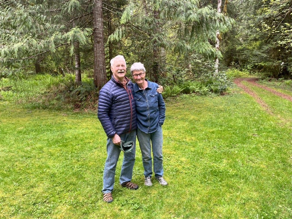 A man and woman with short white hair stand in a green field with trees behind them. Both are smiling. The man has his arm around the woman's shoulders.
