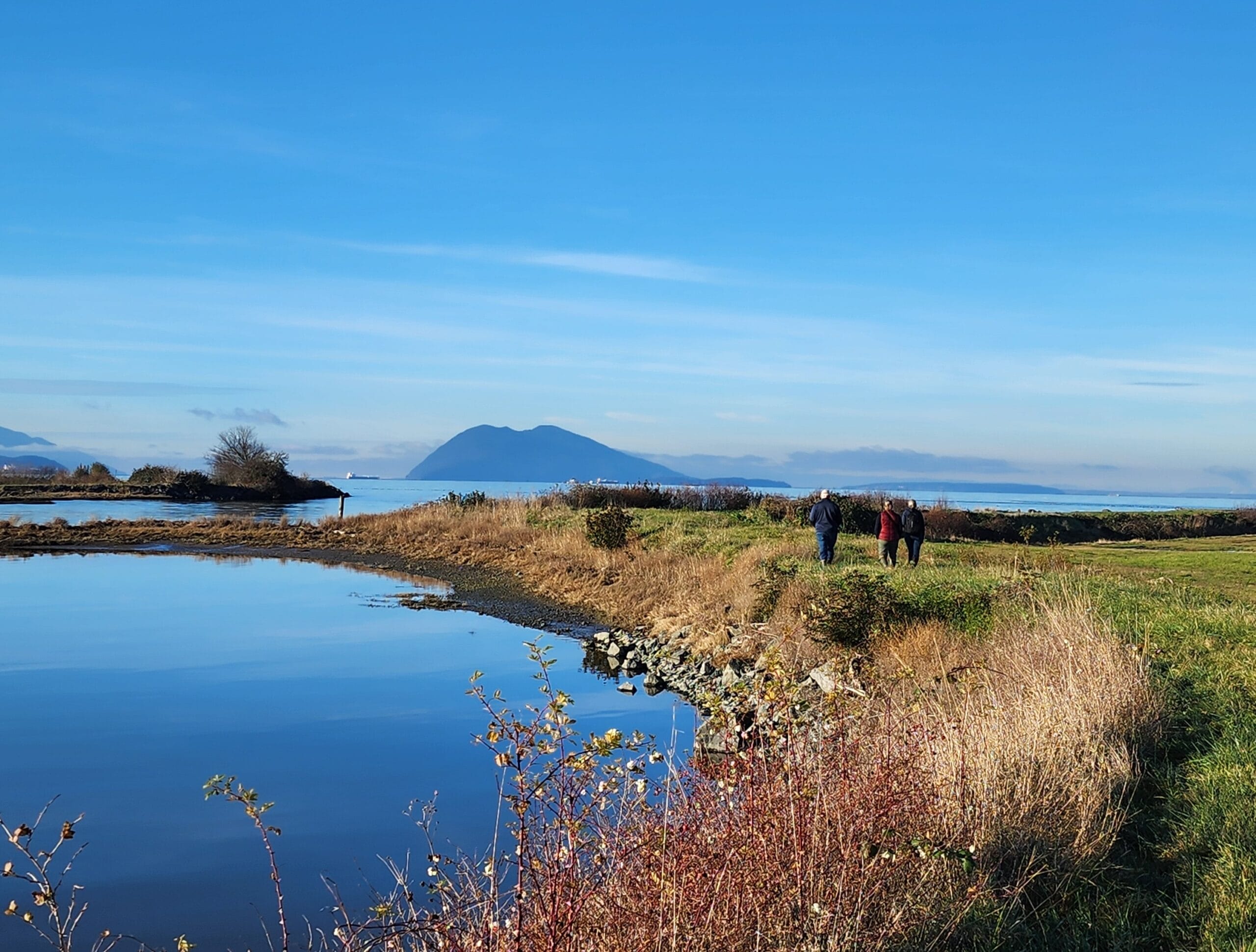 A finger of grassy land curves out into vivid blue water. A vivid blue sky and islands appear in the background. A small group of people walks away from the camera, out onto the finger of land.